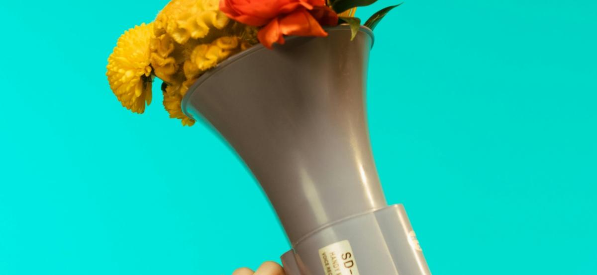 Ein von einer Hand gehaltenes graues Megafon vor türkisem Hintergrund, aus dem ein Blumenstrauß quillt