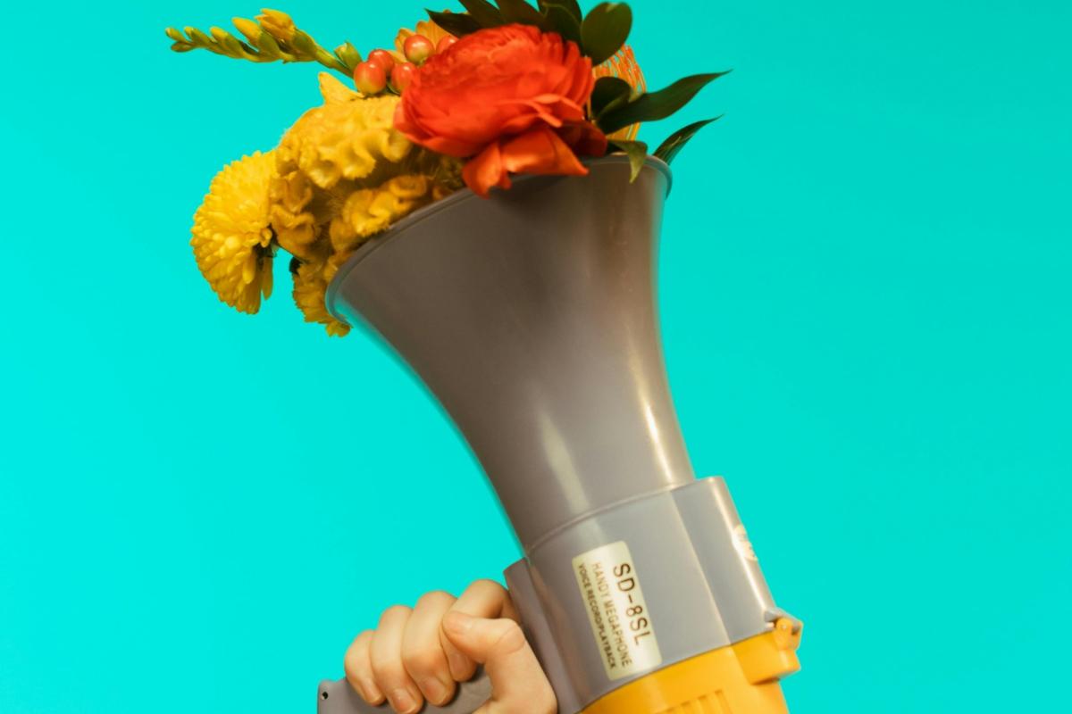 Ein von einer Hand gehaltenes graues Megafon vor türkisem Hintergrund, aus dem ein Blumenstrauß quillt