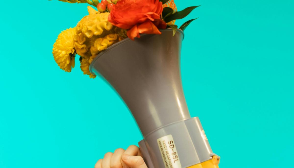 Ein von einer Hand gehaltenes graues Megafon vor türkisem Hintergrund, aus dem ein Blumenstrauß quillt